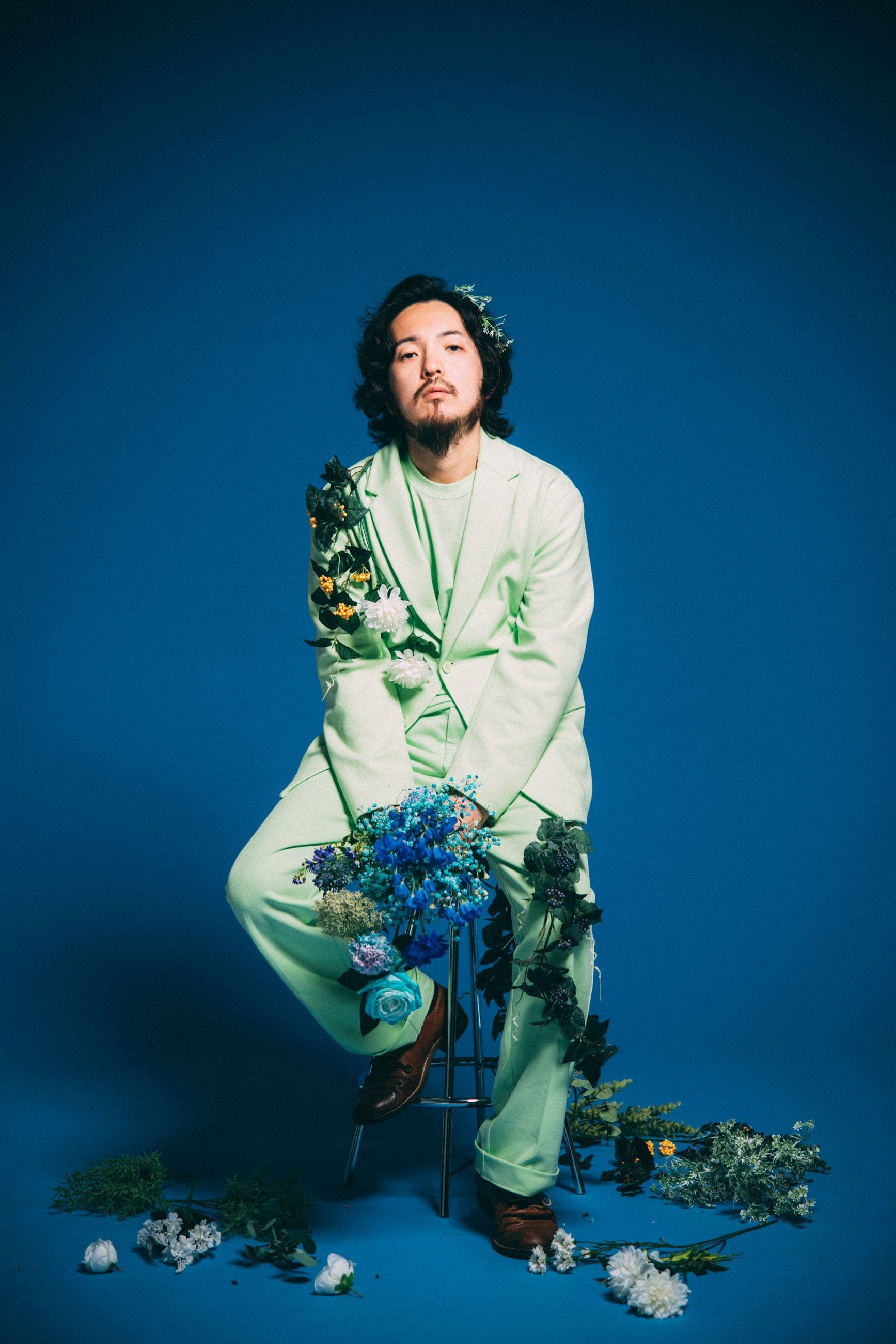 Potomelli seated on a stool in the blue studio, a flower crown and a bouquet of blue flowers resting on his lap.